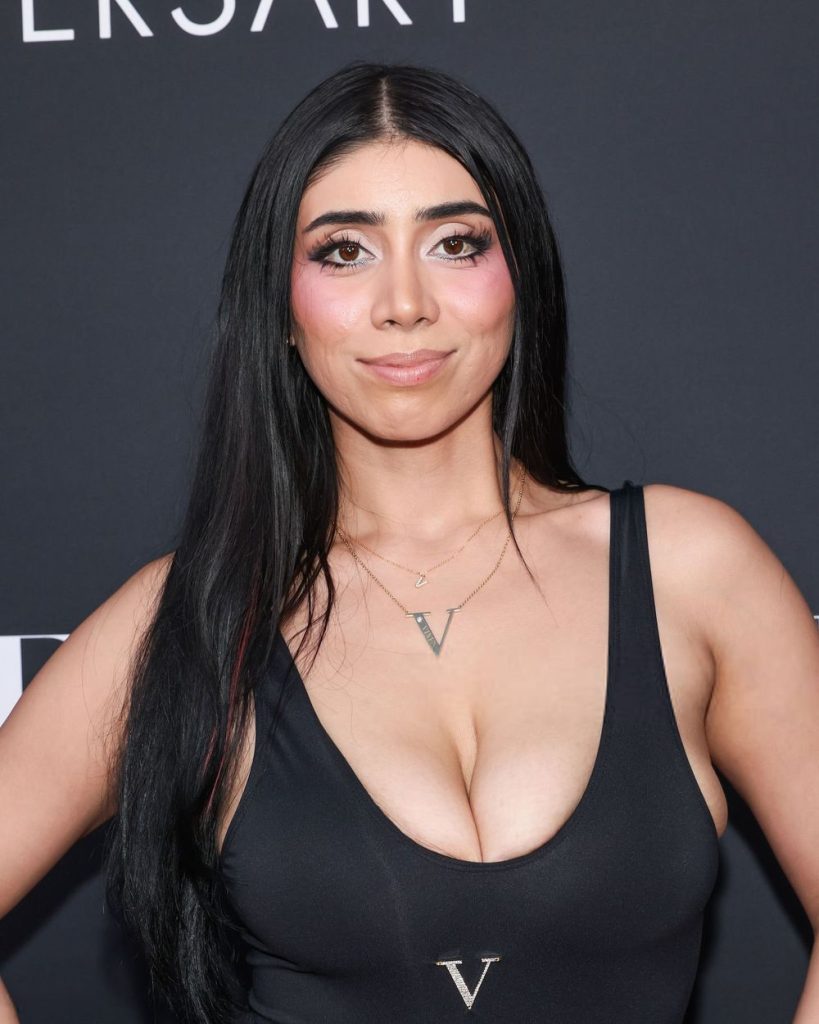 A woman with long black hair, wearing a black sleeveless top and a V necklace, poses in front of a dark background with white text. She has bold makeup and a neutral expression.