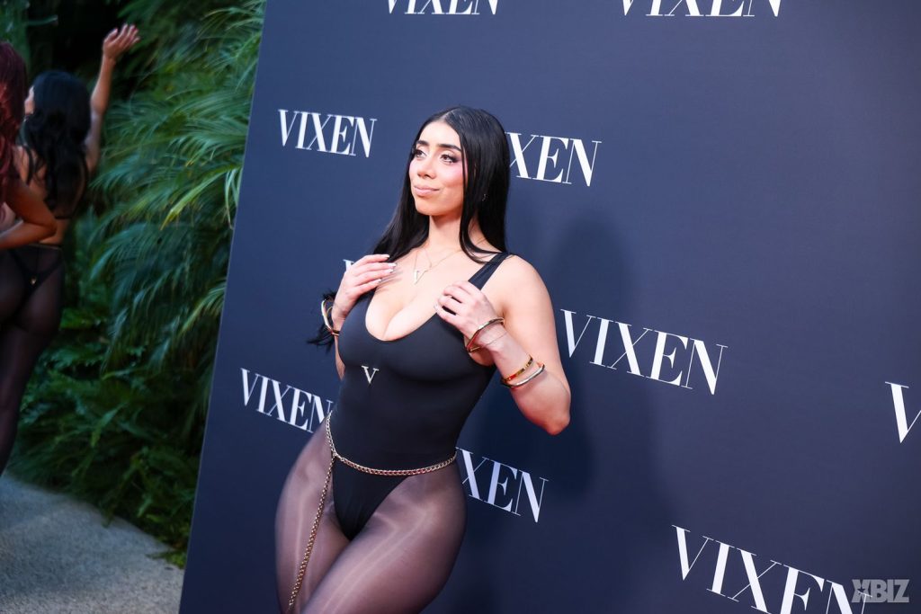 A woman in a sheer black bodysuit and gold chain poses in front of a VIXEN branded step-and-repeat backdrop at an event. Lush greenery is visible to the left.