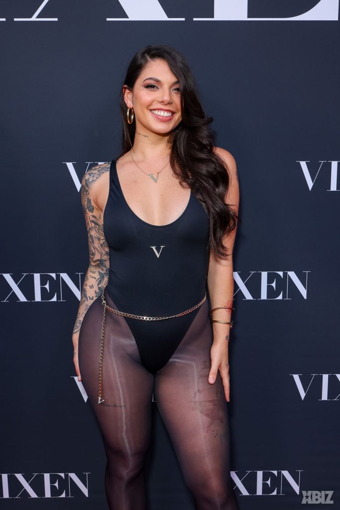 A woman with long dark hair wearing a black bodysuit and sheer tights poses and smiles in front of a backdrop with the word VIXEN and the XBIZ logo. She has tattoos on her arm and is accessorized with earrings and a chain belt.