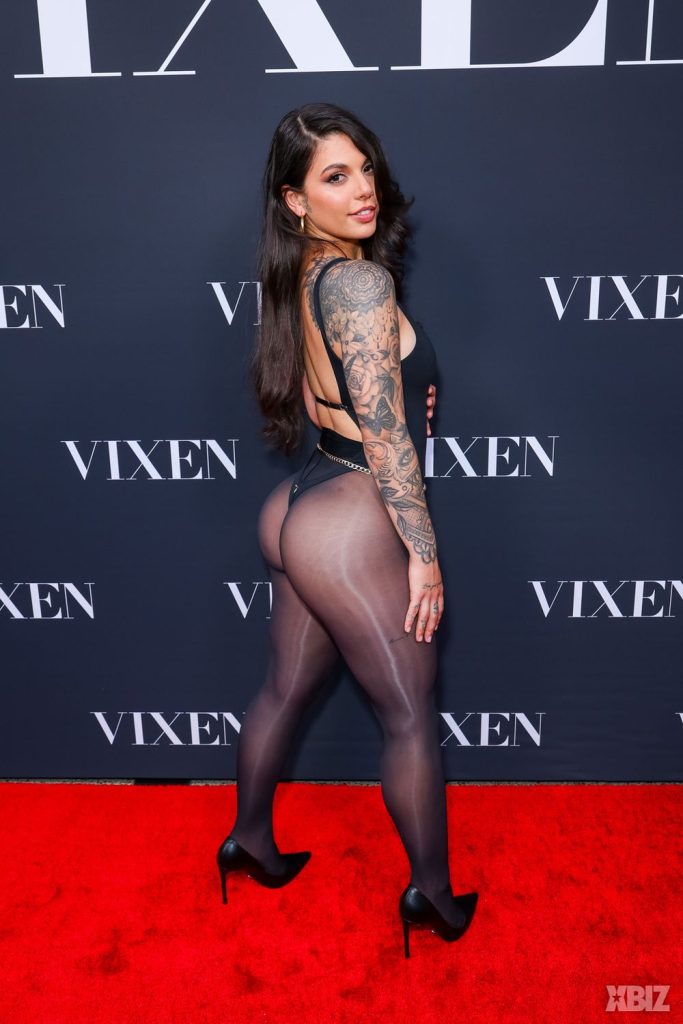 A woman with long dark hair and a full arm tattoo poses on a red carpet in a black bodysuit and heels, standing in front of a backdrop with the word VIXEN repeated on it.