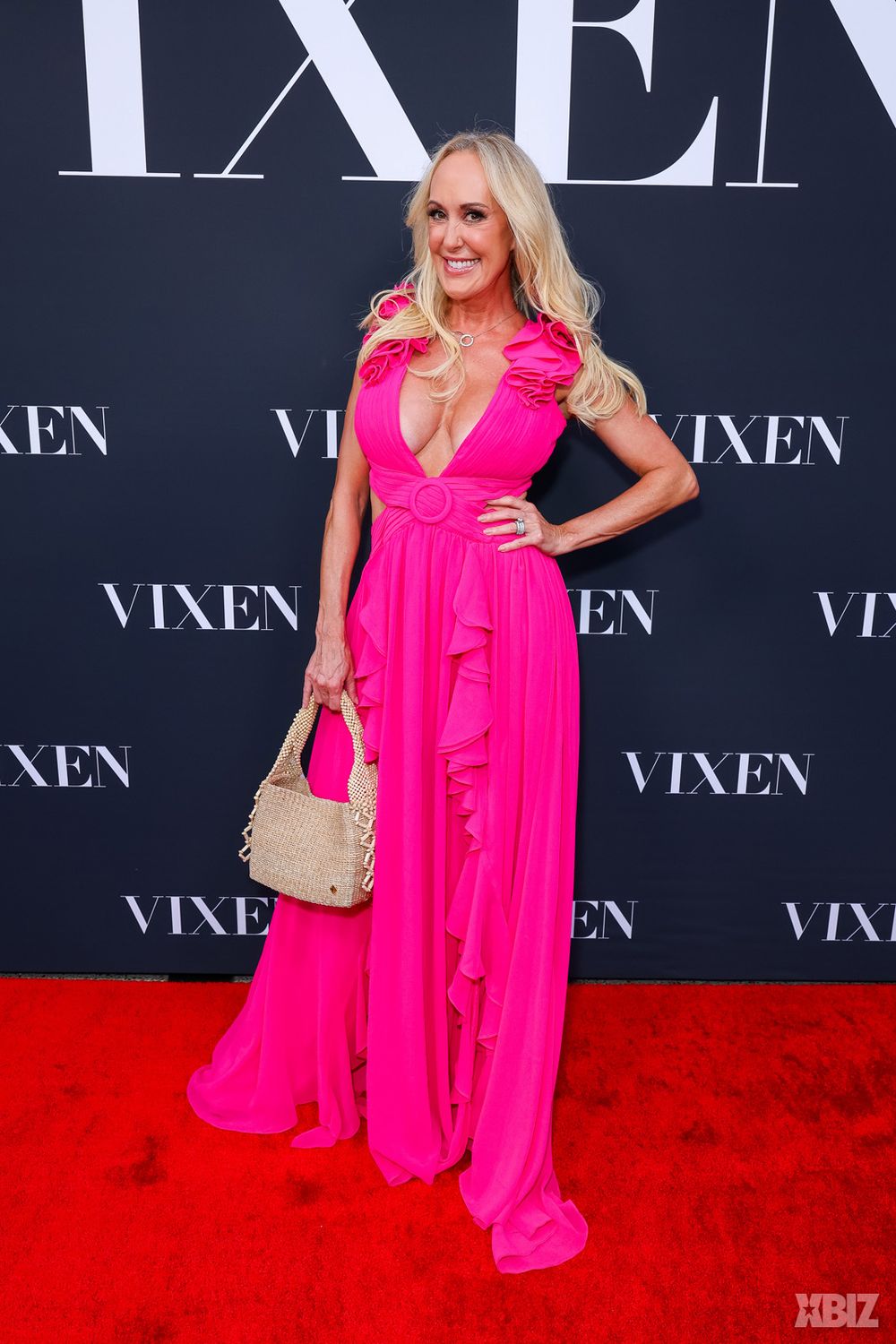 Woman in a bright pink ruffled gown poses on a red carpet, smiling and holding a textured beige handbag, with a black backdrop displaying the word VIXEN in bold white letters.