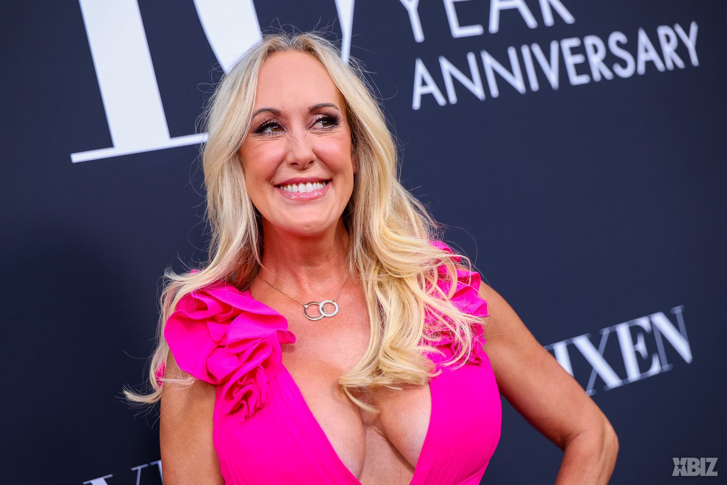 A smiling woman with long blonde hair wears a pink dress with ruffled sleeves, posing in front of a black backdrop with white anniversary text.