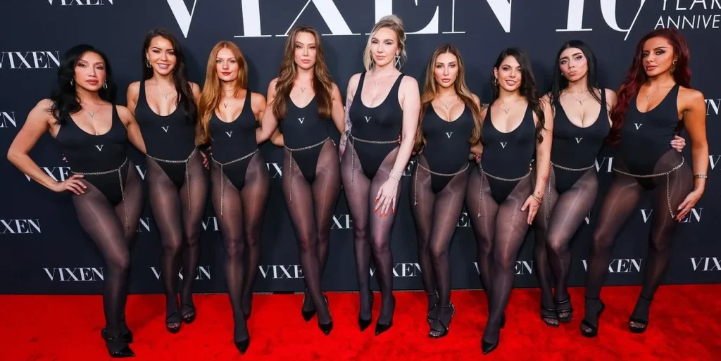Nine women in matching black bodysuits and sheer black tights pose together on a red carpet in front of a VIXEN 10 Year Anniversary backdrop.