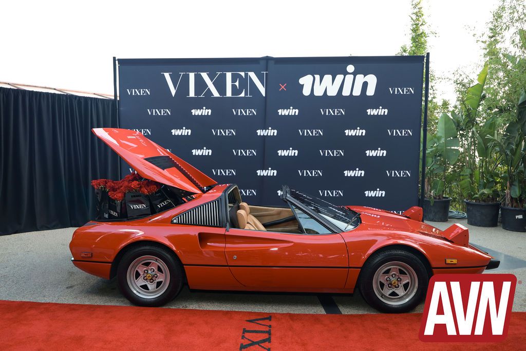 A red convertible sports car with the trunk open is displayed on a red carpet in front of a black VIXEN x 1win backdrop. Large AVN logo in the bottom right corner.