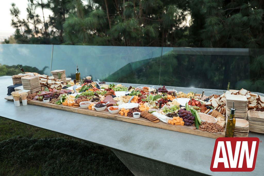 A long outdoor table with assorted cheeses, fruits, sandwiches, crackers, and dips, set against a backdrop of green trees. The AVN logo appears in the lower right corner.