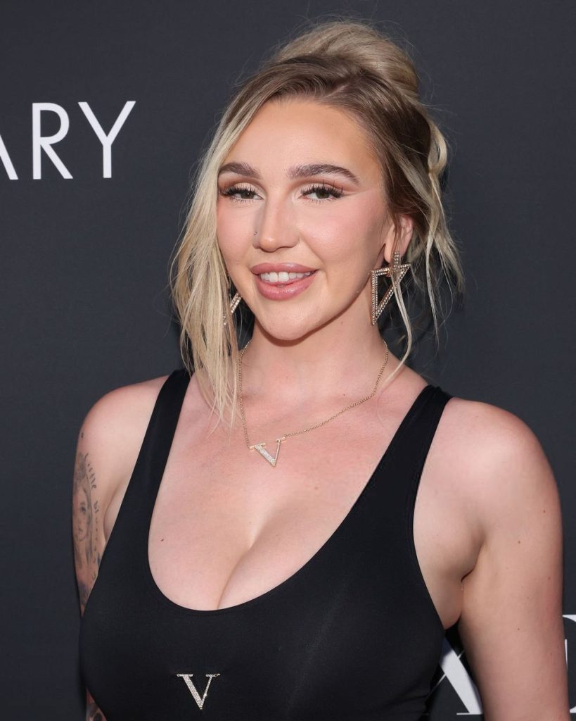 A woman with blonde hair in an updo, wearing a black top, V-shaped necklace, and geometric earrings, smiles in front of a dark backdrop with partial white lettering.
