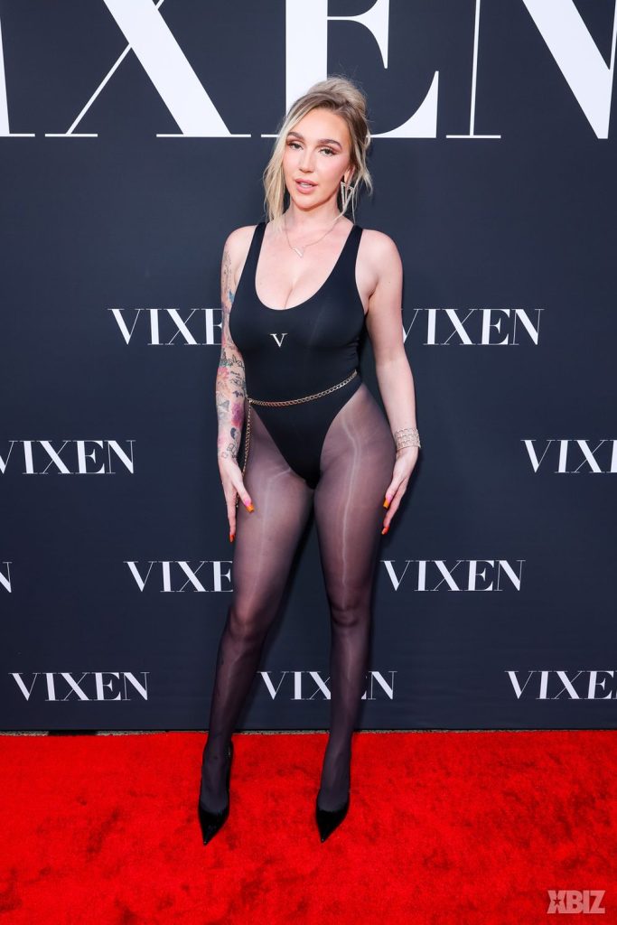 A woman poses on a red carpet in a black bodysuit and sheer black tights, wearing black heels. The background features the word VIXEN in large white letters.