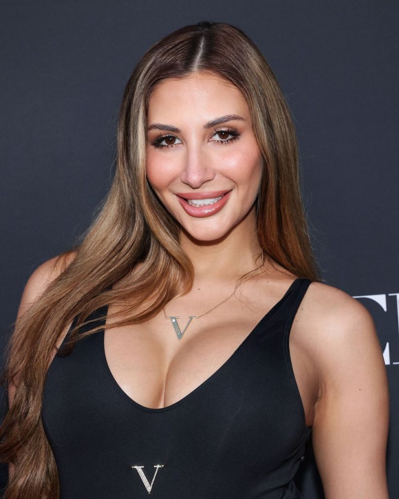 A woman with long brown hair in a black sleeveless top smiles at the camera against a dark background. She wears a necklace with a V pendant.