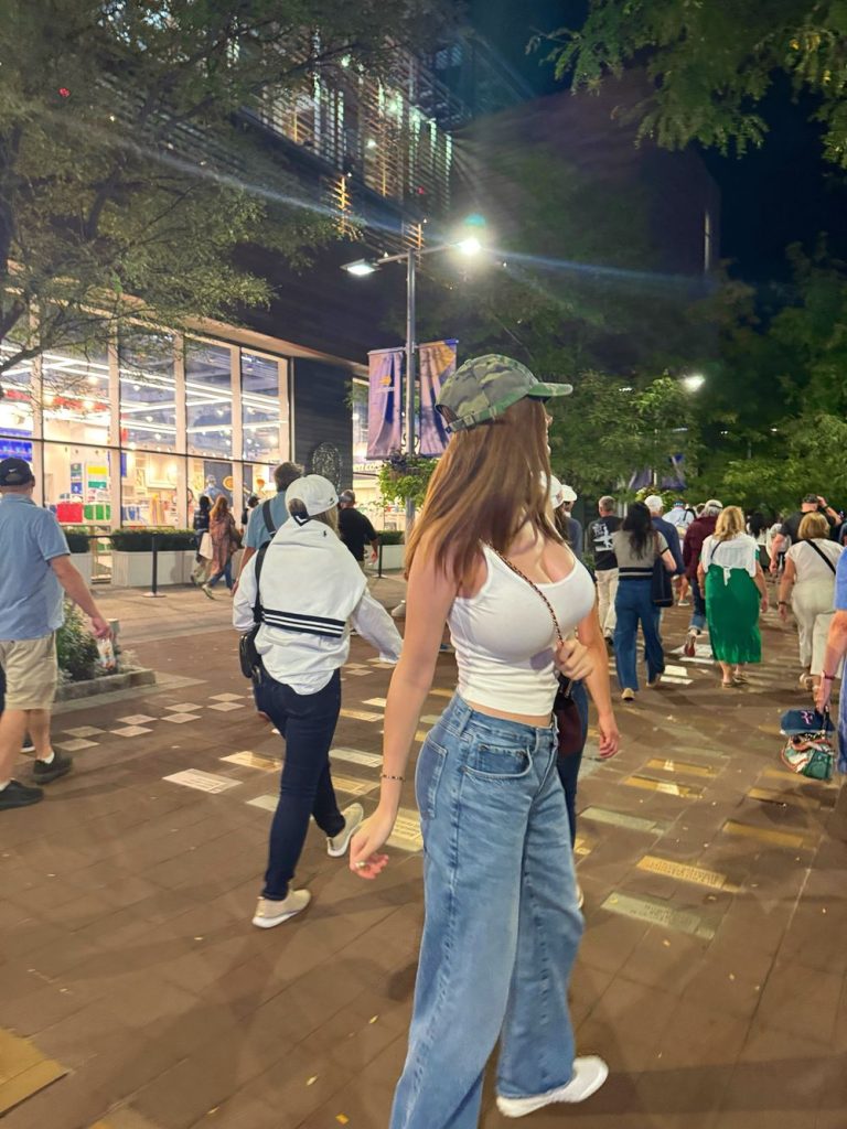 A woman in a white tank top, jeans, and a camouflage cap walks on a busy, well-lit city street at night, surrounded by other pedestrians under trees and streetlights.