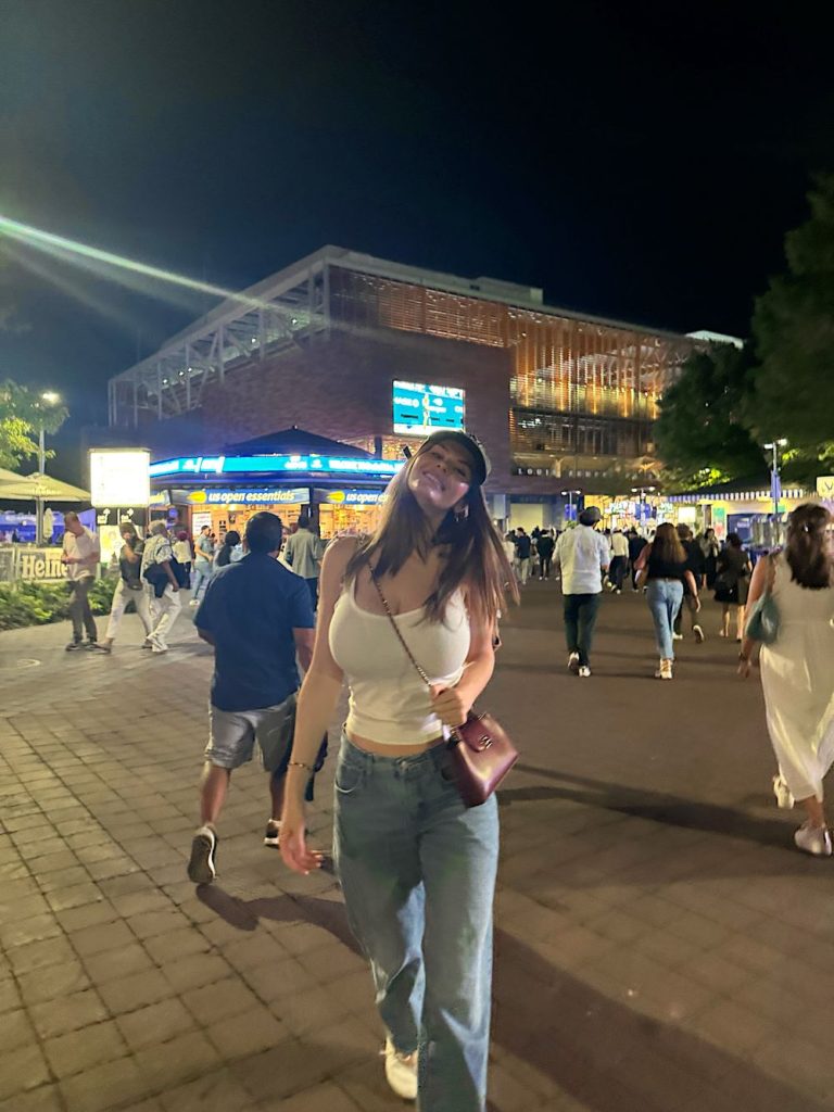 A woman in a white tank top and jeans walks outside at night among a crowd. She is smiling, wearing a cap, and carrying a crossbody bag. A brightly lit building is in the background.