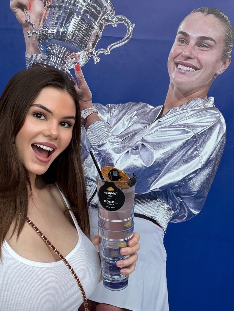 A smiling woman holds stacked plastic cups and poses in front of a large poster of a female tennis champion holding a trophy.