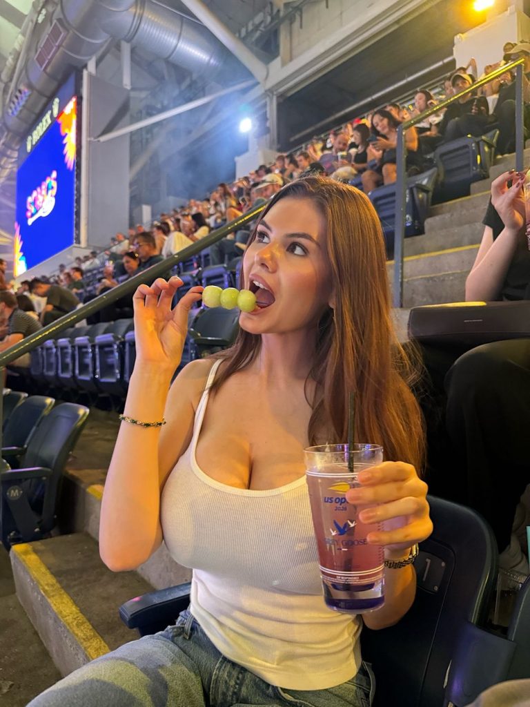 A woman in a white tank top sits in stadium seats, eating grapes from a skewer and holding a drink. The stands are filled with people and a scoreboard is visible in the background.