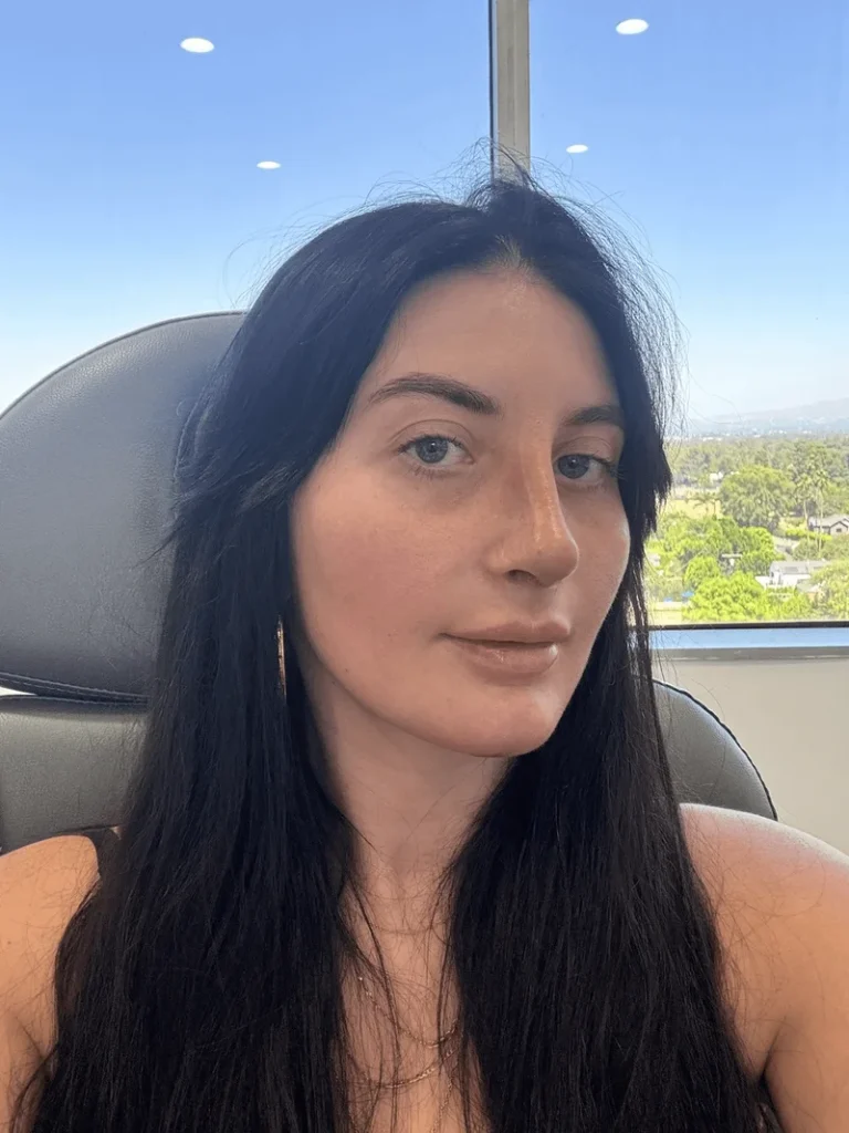 A woman with long dark hair takes a selfie indoors, seated in front of a large window with a view of trees and a clear blue sky outside.