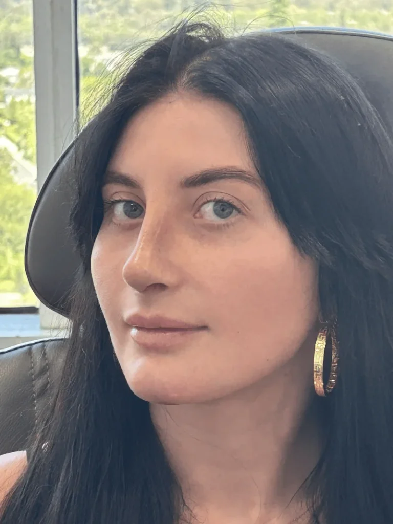 A woman with long dark hair and light eyes, wearing gold hoop earrings, sits in a chair in front of a window with greenery visible outside.