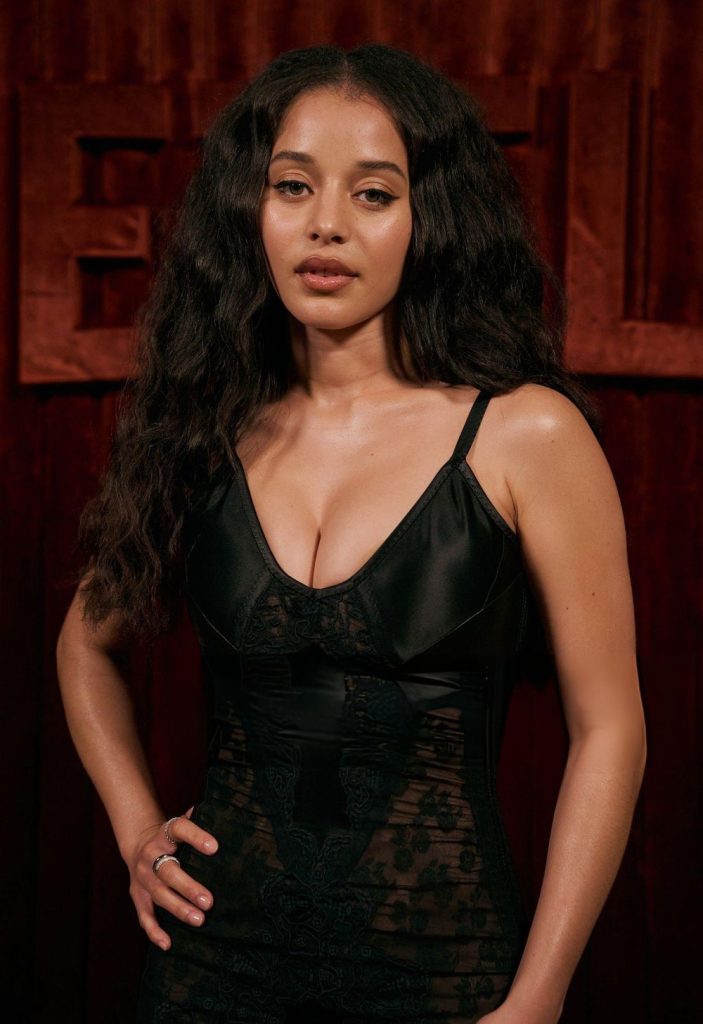 A woman with long, wavy dark hair wears a fitted black dress with lace details, standing confidently in front of a dark red background with large letters.