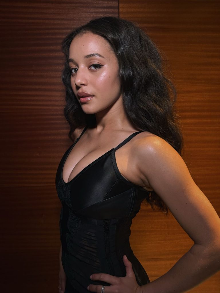 A woman with long wavy hair wearing a black sleeveless top stands in front of a wooden background, looking confidently at the camera.