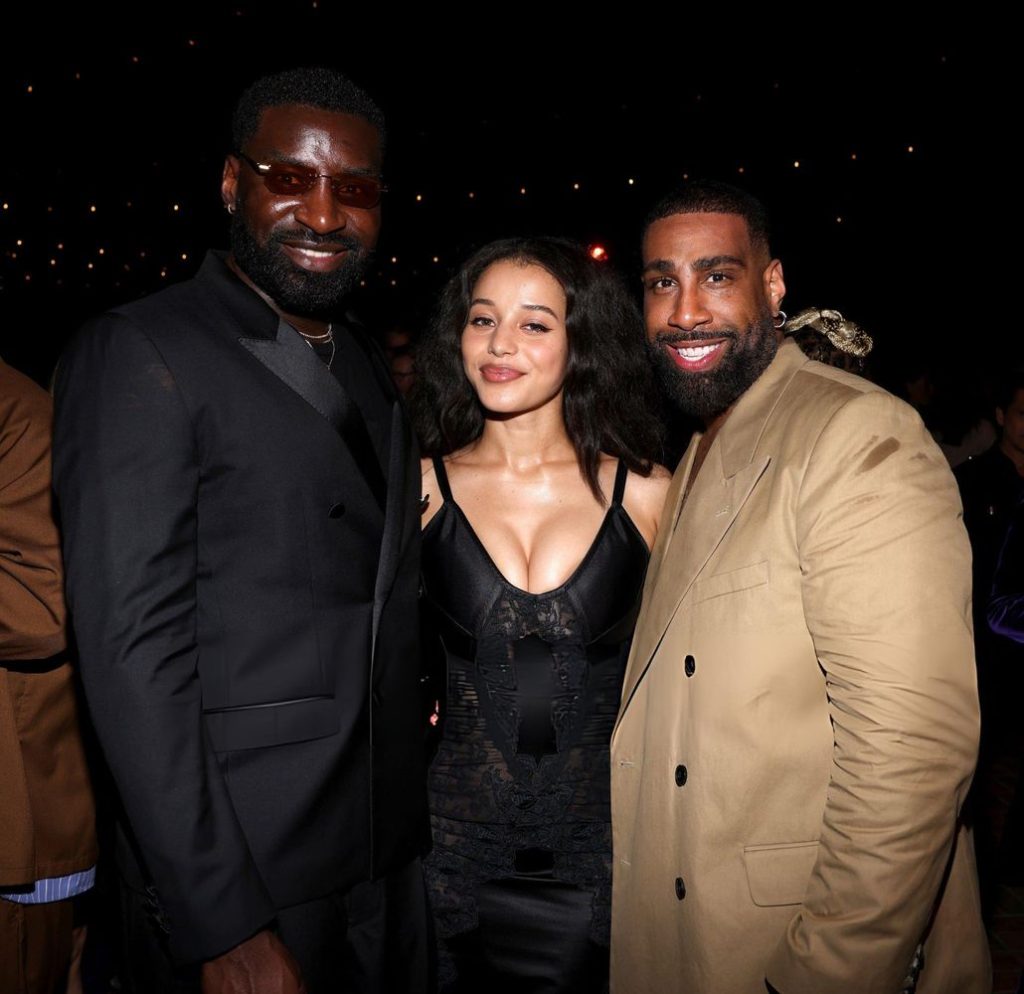 Three people pose together at a nighttime event. The man on the left wears a black suit, the woman in the middle wears a black dress, and the man on the right wears a tan coat. All are smiling.