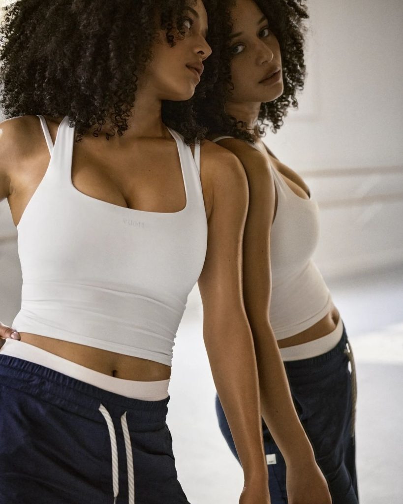 A woman with curly hair wears a white tank top and navy blue pants, leaning against a mirror and gazing at her reflection in a softly lit room.