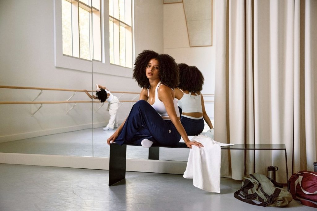A woman in workout clothes sits on a bench near a towel and gym bag in a dance studio with mirrors, while another person dances in the background.