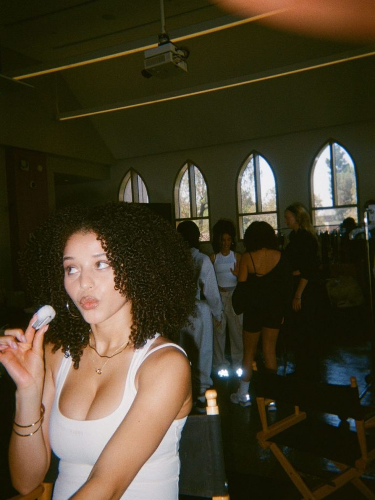 A woman with curly hair in a white tank top holds makeup, looking sideways, in a room with arched windows and several people in the background.