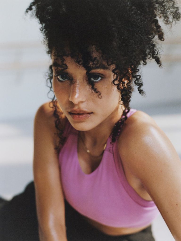 A person with curly hair looks intently at the camera, wearing a sleeveless pink top. Their skin is slightly glistening, suggesting they may have been exercising. The background is softly blurred.