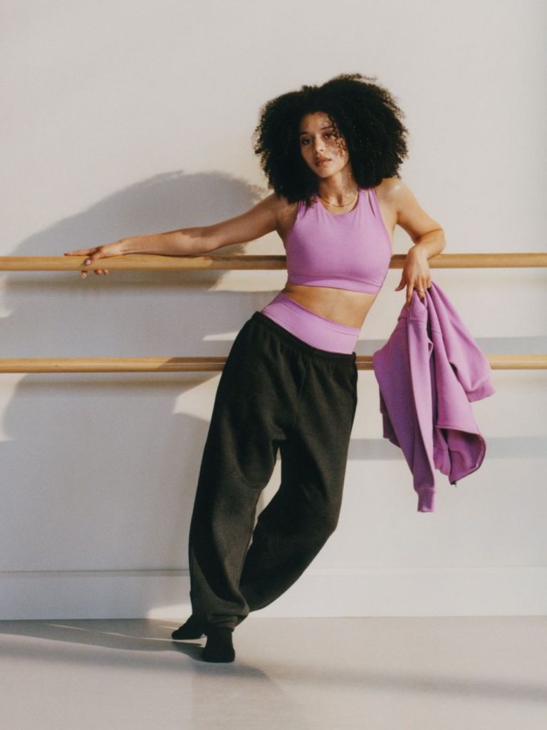 A woman in a purple crop top and black sweatpants leans on a ballet barre, holding a matching purple jacket. She stands barefoot in a bright room with white walls and wooden barres.