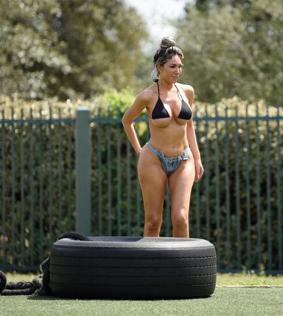 Woman with blonde-highlighted hair in a bun outdoors on artificial turf, wearing a black triangle bikini top and denim thong bottoms, standing inside a large black tire with one hand on her hip.