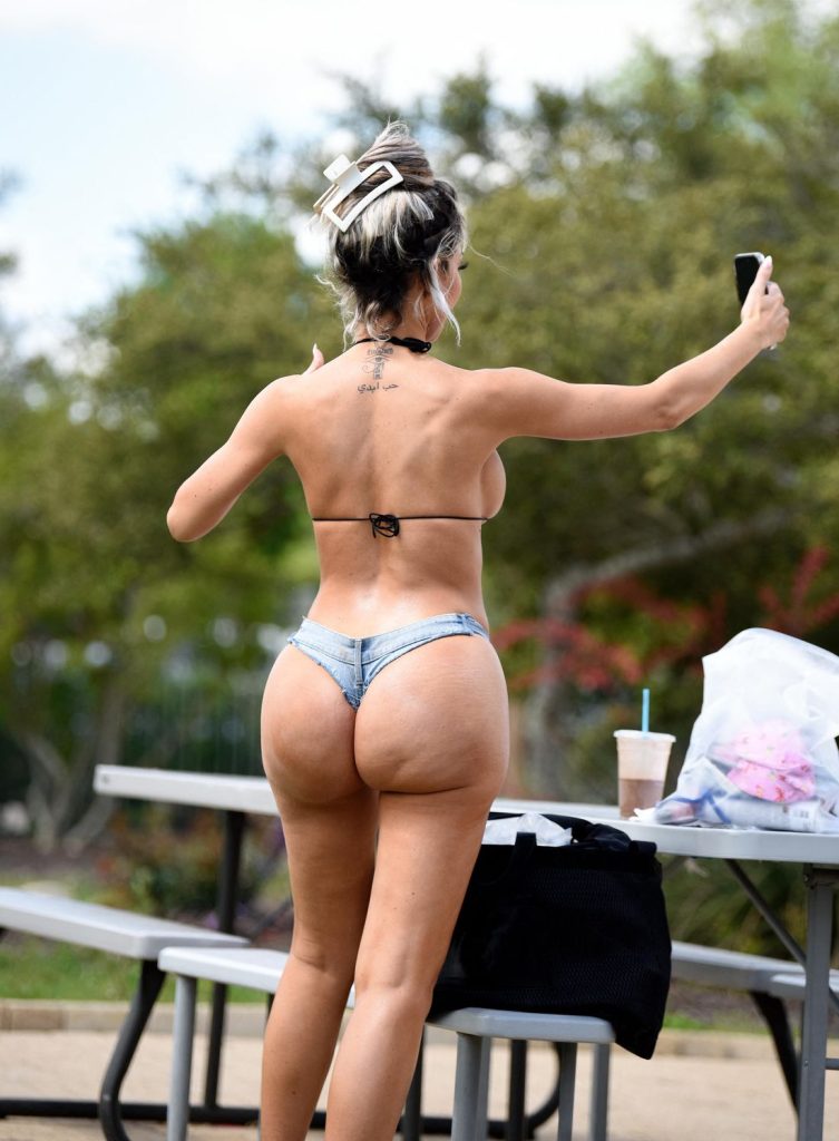 Woman with blonde-highlighted hair in a bun outdoors, wearing a black triangle bikini top and denim thong bottoms, viewed from behind while holding a phone up near a picnic table.