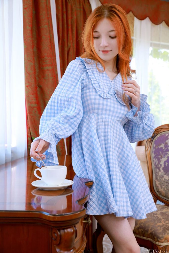 Red-haired woman standing beside a wooden desk, looking down while stirring a white teacup with a spoon, wearing a light blue gingham dress.