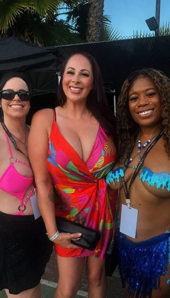 Three women smiling outdoors. One wears a pink bikini top and black skirt, another wears a colorful dress, and the third wears a blue beaded top and skirt. Trees and a tent are visible in the background.