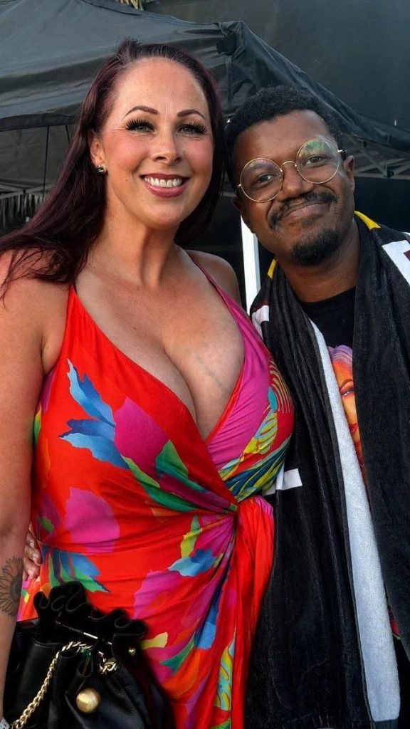 Two people smiling and posing together outdoors. The woman wears a colorful floral dress and carries a black purse. The man wears glasses, a dark shirt, and a towel draped over his shoulders. Theres a tent structure in the background.
