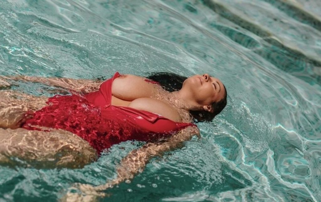 Woman with dark wet hair floating on her back in a swimming pool, wearing a red one-piece swimsuit that is partially open at the chest, eyes closed.