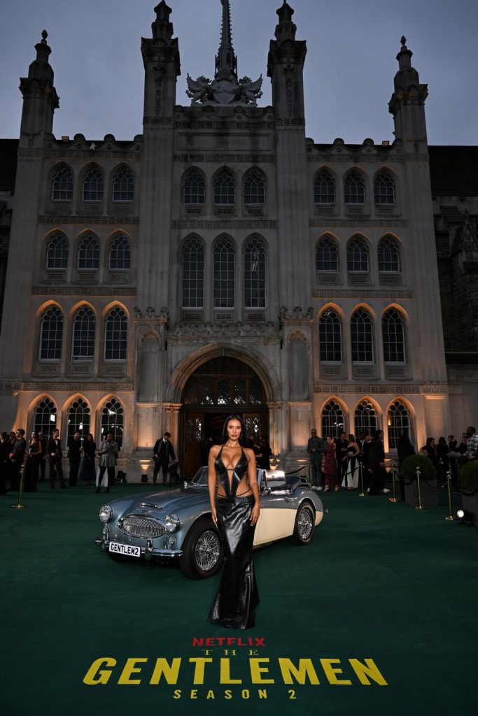 A woman in a dramatic black dress stands in front of a classic car at dusk, outside a grand historic building, with The Gentlemen Season 2 and Netflix branding on a green carpet.