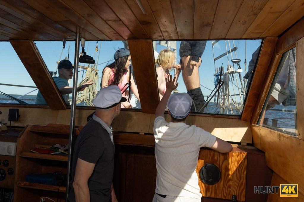 View from inside a boat cabin looking out a window at a blonde woman in a bikini performing oral sex on a man on the deck, with others watching.