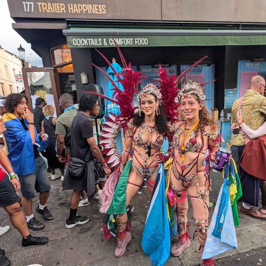 Two women in elaborate, feathered carnival costumes stand smiling on a city street, surrounded by people and colorful buildings, in front of a place called Trailer Happiness..