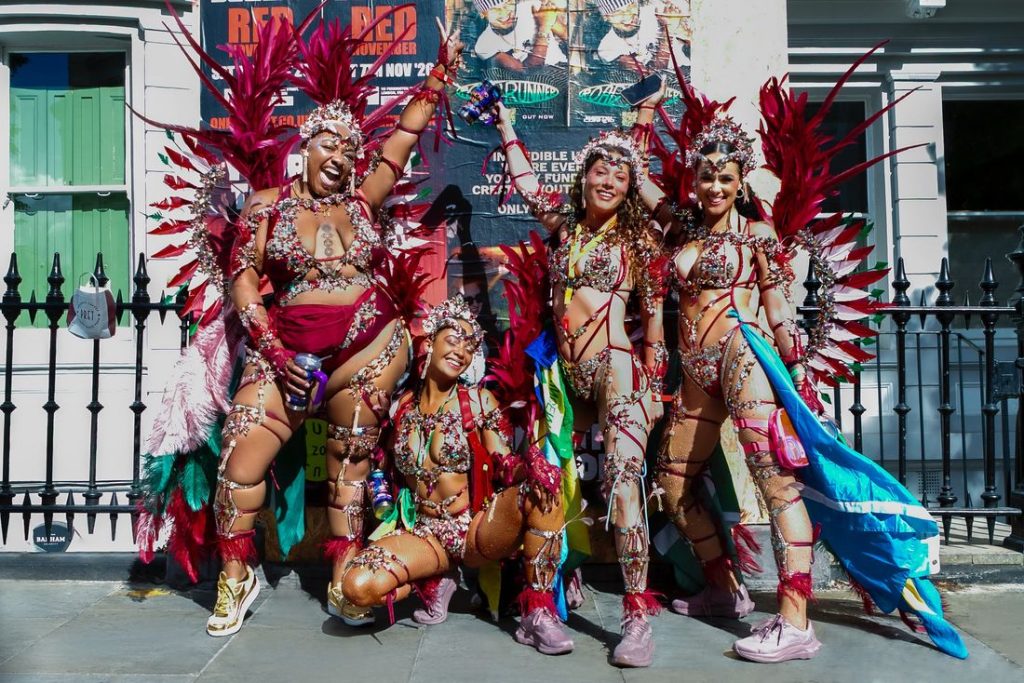 Four people in vibrant, feathered carnival costumes pose and smile energetically on a sunny street, with colorful wings, beaded outfits, and festive accessories in front of a fence and posters.
