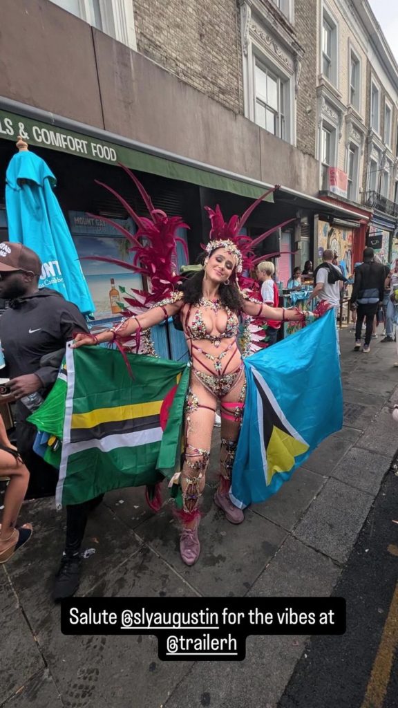 A woman in a colorful, feathered carnival costume holds flags of Dominica and the Bahamas on a busy city street, surrounded by people and shops.