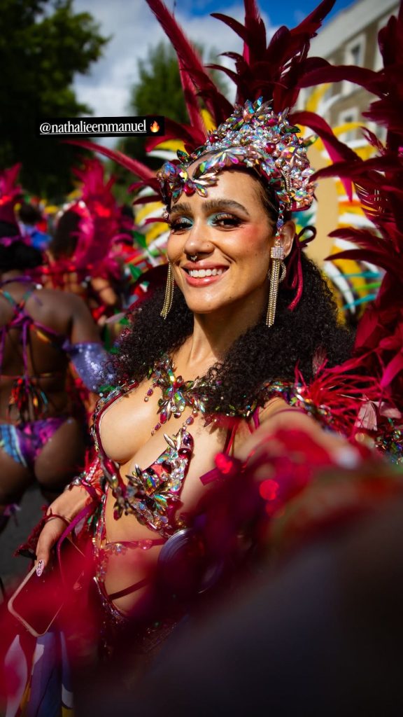 A woman in a vibrant, jewel-encrusted carnival costume with red feathers smiles at a parade. She has curly hair, bold makeup, and decorative headpiece, surrounded by others in colorful attire.