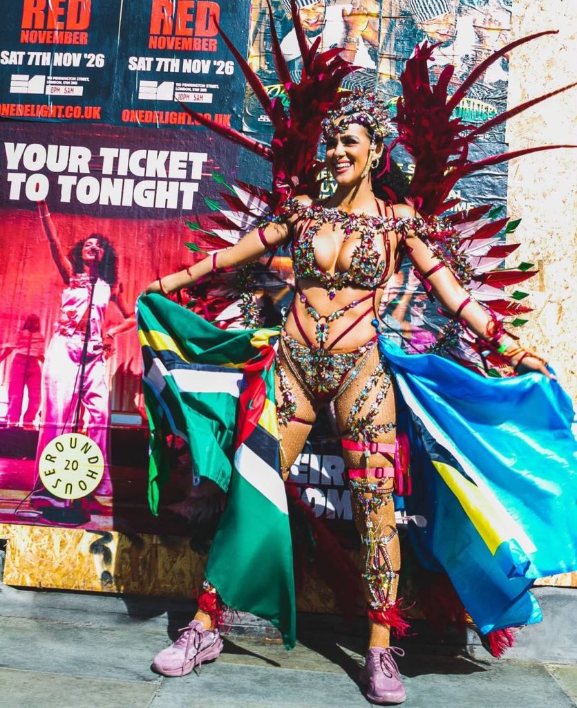A woman in a colorful, feathered carnival costume poses outdoors, holding flags. Posters and wooden boards are visible behind her.