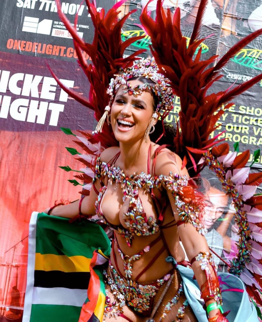 A smiling woman in a colorful, gem-encrusted carnival costume with large red feathered wings holds a flag. She stands in front of a vibrant event poster background.
