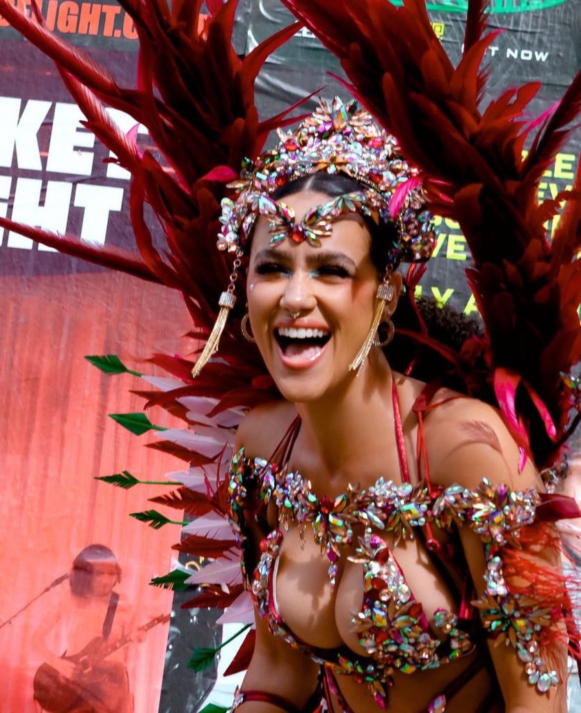 A woman in a colorful, bejeweled carnival costume with large red feathered wings smiles and laughs at an outdoor event.