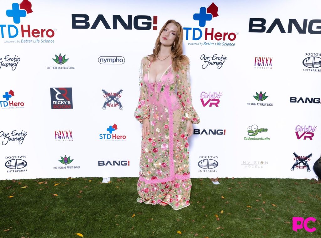 A woman in a sheer floral dress with pink accents poses on grass in front of a sponsor step-and-repeat backdrop at an outdoor event.