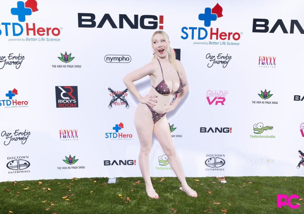 A woman poses outdoors on grass in a bikini, standing in front of a backdrop with multiple event sponsor logos.