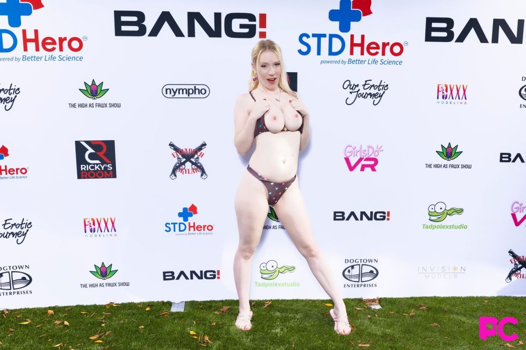 A fair-skinned woman with blonde hair wearing a brown, polka-dot bikini poses in front of a white step-and-repeat backdrop featuring various company logos, standing on a patch of artificial green grass. She is topless.