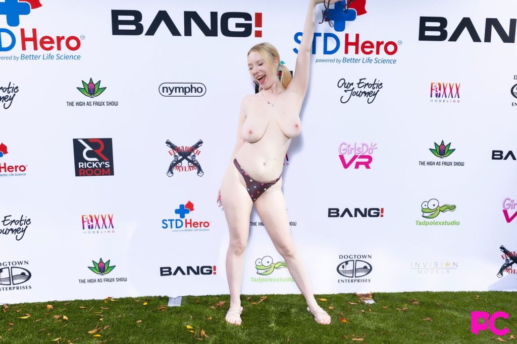 A topless woman posing confidently in a swimsuit at an outdoor event, standing in front of a backdrop featuring various sponsor logos on the grass.