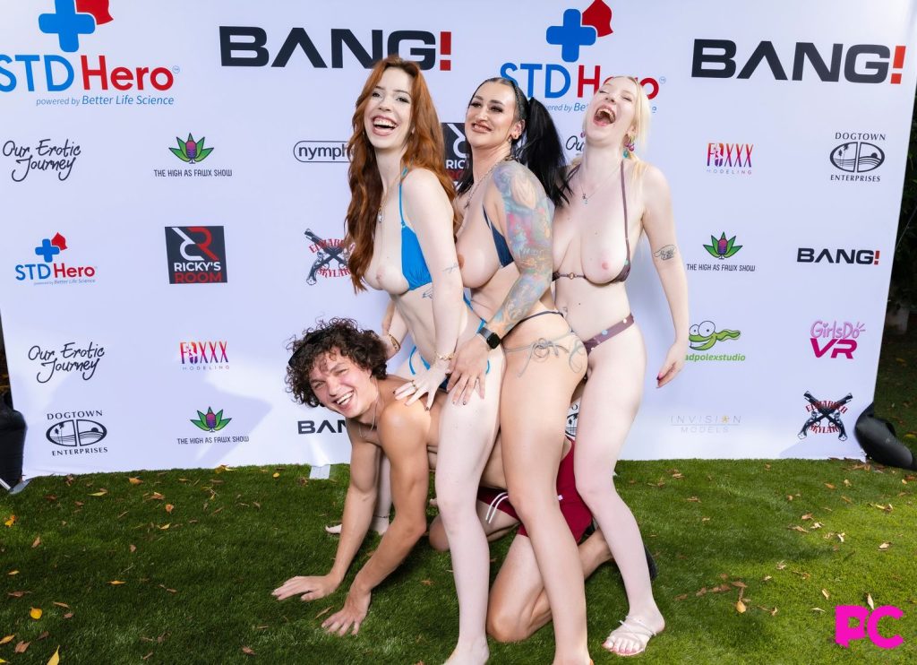Four people in swimwear pose playfully outdoors on grass, with three women kneeling or standing on a smiling man, in front of a promotional event backdrop with various sponsor logos.
