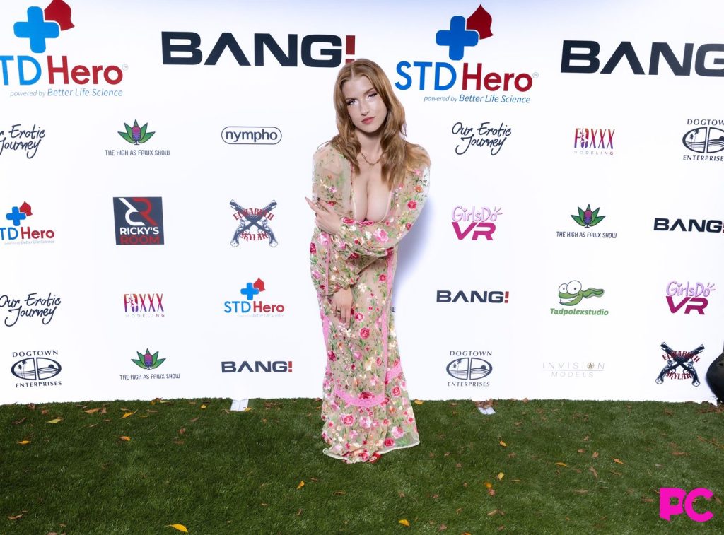 A woman in a floral dress poses, arms crossed, on a grass carpet in front of a sponsor logo backdrop at an event.