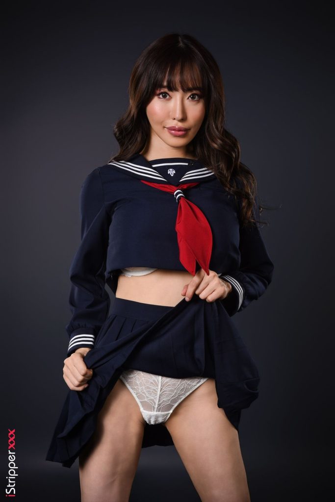 A young Asian woman with long brown hair, wearing a dark navy blue sailor-style school uniform with a red neckerchief, lifting the skirt to reveal white lace lingerie against a solid dark grey studio background.