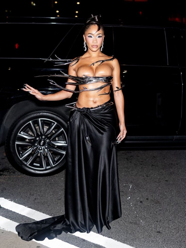 A woman poses on the street at night, wearing a dramatic black outfit with metallic strips across her chest and a long silk skirt, standing in front of a dark vehicle.
