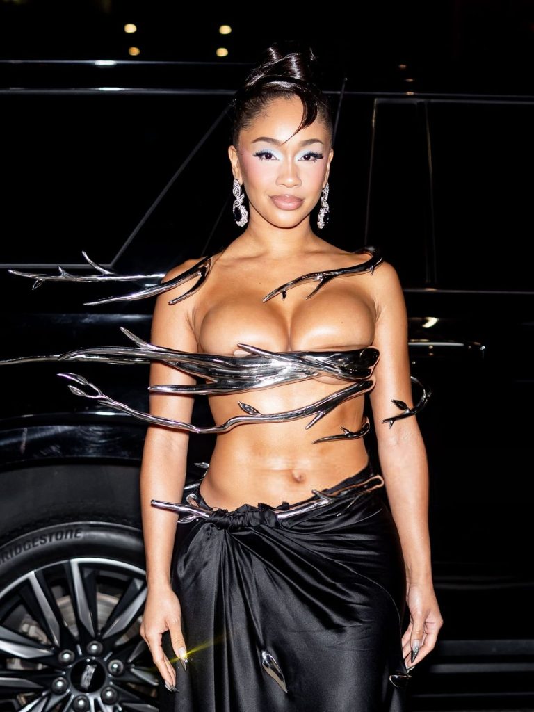 A woman poses in front of a black car, wearing a striking black outfit with metallic, branch-like details wrapping around her upper body and a draped black skirt. She has statement earrings and an elegant updo hairstyle.